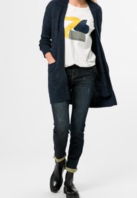 Cardigan en maille bleu marine à ouverture frontale, t-shirt graphique blanc avec des formes abstraites en jaune et bleu, jean foncé et bottines noires.
