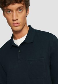 Polo shirt blu navy scuro realizzato in tessuto morbido; presenta una tasca sul petto, una patta con bottoni e un sottile logo decorativo sulla tasca.