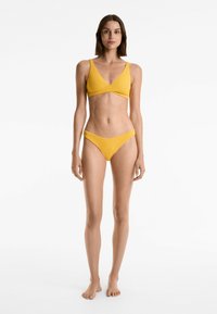 Žlutý žebrovaný bikini set s braletkou ve tvaru V a širokými ramínky, doplněný o sladěné nízké kalhotky. Hladký materiál a pohodlný střih.