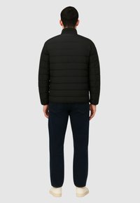 Giacca nera imbottita con design trapuntato orizzontale, colletto alto e zipper frontale. Indossata con jeans scuri e scarpe di colore chiaro.