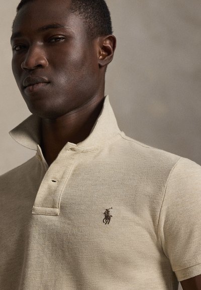 Povečava moškega, ki nosi bež polo majico z dvignjenim ovratnikom in majhnim vezenim logotipom konja in jezdeca na prsih.