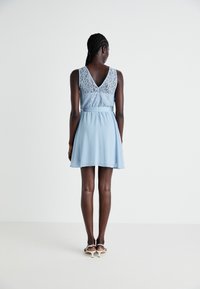 Svetlo modra obleka z čipkastim korzetom in V-izrezom na hrbtni strani, z lahkotno krilo in dolžino do kolen. Material se zdi mehak z gladko teksturo.