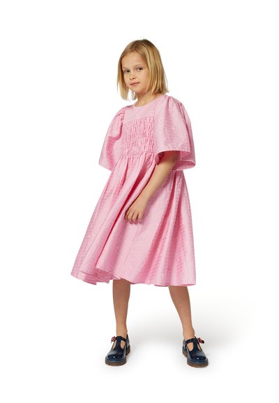 Robe rose avec un motif texturé, corsage froncé et manches volantées. Associée à des chaussures T-bar bleu marine ornées d'une fleur.