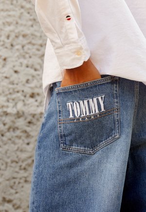 Person trägt blaue Jeans mit "TOMMY JEANS" auf der Gesäßtasche bestickt und ein weißes Langarmshirt.