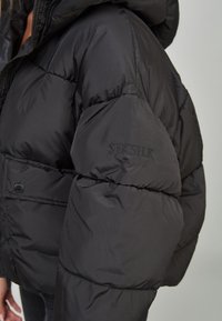 Chaqueta de plumas negra con un diseño corto, textura acolchada y un logotipo bordado en la manga. Incluye capucha y botones de presión.