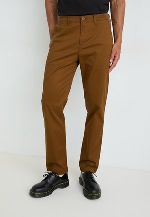 Chinos marrones de corte ajustado, con cierre de botón y trabillas para cinturón, combinados con zapatos negros de cordones y una blusa negra.