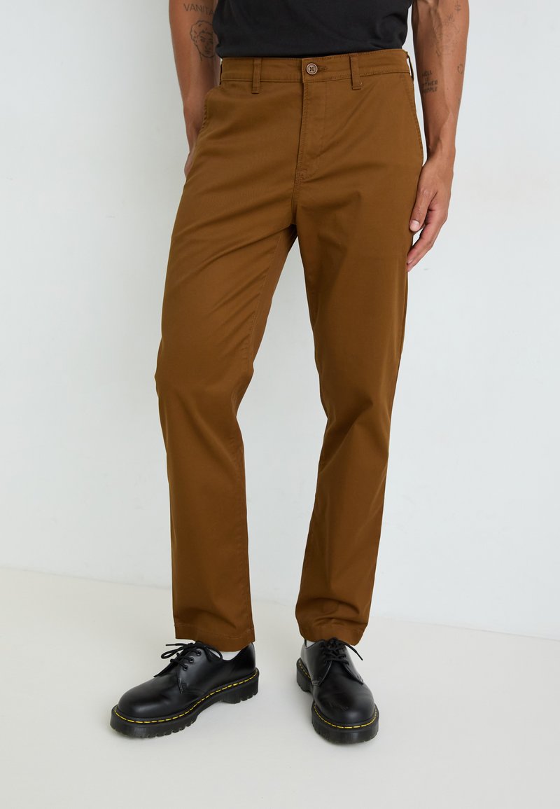 Brown chinos with a slim fit, featuring a button closure and belt loops, paired with black lace-up shoes and a black top.