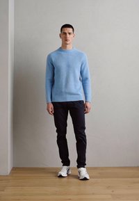 Un uomo è in piedi contro una parete semplice, indossa un maglione blu chiaro, pantaloni neri e sneakers bianche su un pavimento di legno.