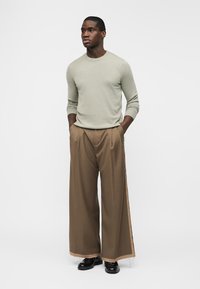 Man draagt een beige shirt met lange mouwen, wijd uitlopende bruine broek met beige bies, zwarte schoenen, staat met handen in de zakken tegen een witte achtergrond.