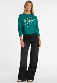 Mlada ženska v turkiznem sweatshirtu z napisom "guess jeans" in črnimi širokimi hlačami, stoji ob beli steni.