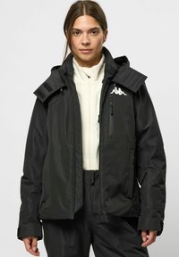 Schwarze wasserdichte Jacke mit hohem Kragen, verstellbarem Kapuze und Reißverschlusstaschen; mit Logo auf der Brust und Klettverschluss-Akzenten an den Schultern.