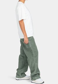 T-shirt blanc à manches courtes associé à un pantalon en velours côtelé vert ample, avec des poches sur les côtés et un tissu texturé. Modèle portant des baskets beiges.