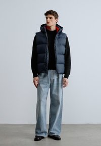 Colete acolchoado azul escuro com capuz, usado sobre um suéter preto. Combinado com jeans de perna larga azul claro e sapatos pretos.