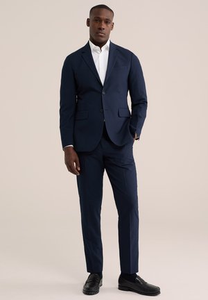Mand iført en mørkeblå skræddersyet habit, hvid skjorte, sorte loafers, der står med den ene hånd i lommen mod en ensfarvet lys baggrund.