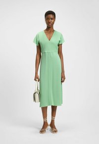 Vihreä midi-mekko V-pääntiellä, lyhyillä hihoilla ja sileällä tekstuurilla. Malli pitää kädessään pientä beigepunosta käsilaukkua ja kantaa ristikkäisiä sandaaleja.
