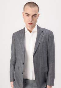 Blazer grigio a righe con due bottoni, rever a punta e tasche laterali, abbinato a una camicia bianca. Tessuto liscio.