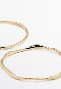 Bracciali in tonalità oro con un design intrecciato, caratterizzati da una texture liscia e una forma irregolare. Due bracciali esposti su uno sfondo bianco.
