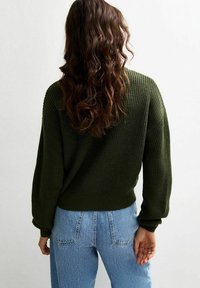 Donkergroene gebreide trui met een losse pasvorm, ribbeltextuur en wijde mouwen, gecombineerd met lichtblauwe high-waisted jeans.