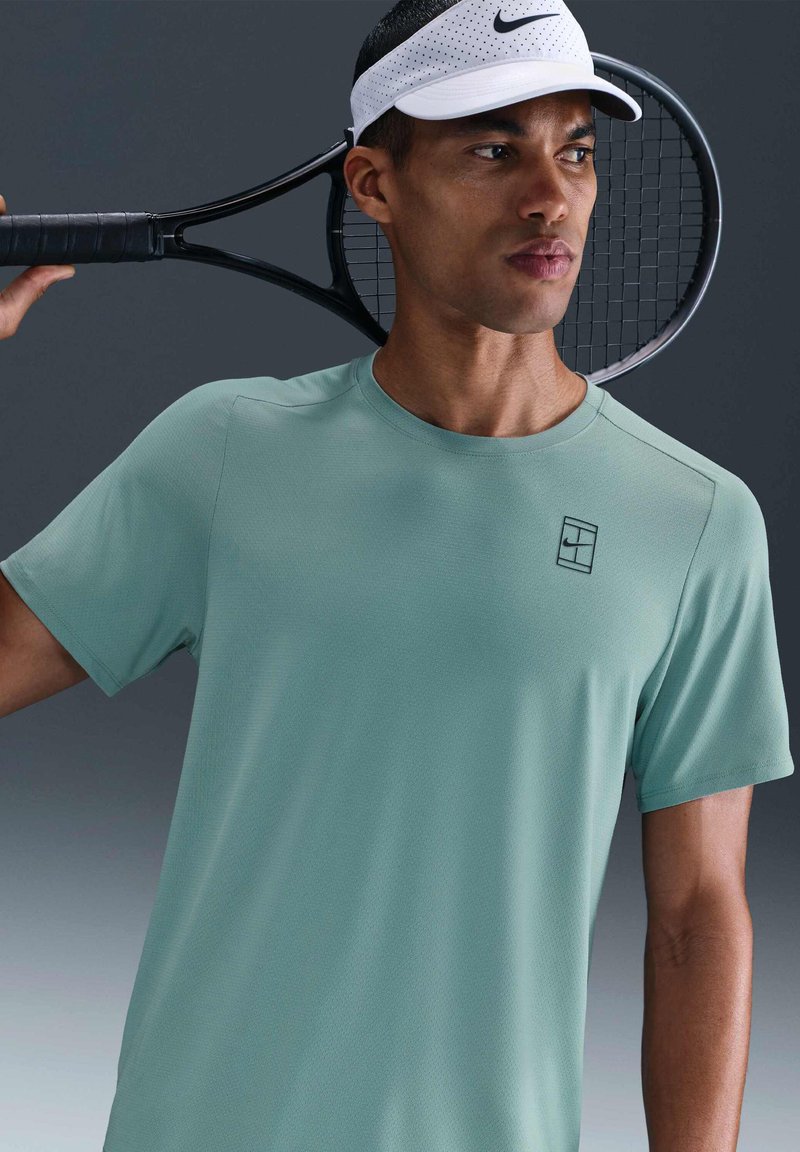 Man in een lichtgroen sportshirt en witte Nike-zonnehoed houdt een zwarte tennisracket over zijn schouder tegen een grijze achtergrond.