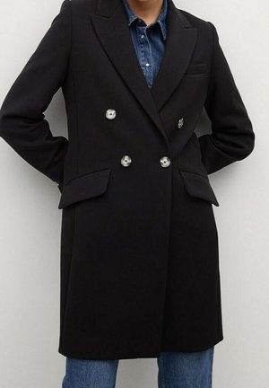 Personne portant un manteau noir à double boutonnage avec des boutons métalliques sur une chemise en jean bleue et un jean bleu, debout les mains dans le dos.