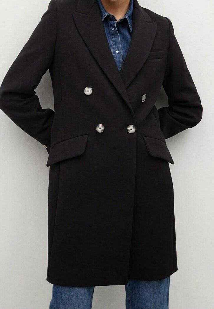 Personne portant un manteau noir à double boutonnage avec des boutons métalliques sur une chemise en jean bleue et un jean bleu, debout les mains dans le dos.