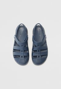 Námořnické modré sandály vyrobené z flexibilního materiálu. Mají otevřený design, pásek přes vrch a texturovanou stélku s odtokovými otvory.