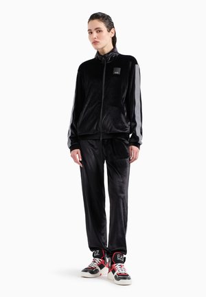 Tuta in velour nero con giacca con zip che presenta un piccolo logo, dettagli a righe sulle maniche e pantaloni abbinati. Indossato con scarpe alte.