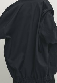 Dunkelblaues, übergroßes Jackett mit rundem Halsausschnitt, elastischem Saum und glattem Stoff mit minimalen Details. Knöpfe an der Seite sichtbar.