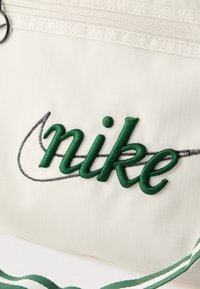 Τσάντα Nike σε ανοιχτό κρεμ ύφασμα με διακριτική υφή, με πράσινο κέντημα λογότυπο, μαύρα ραφές και ριγωτό λουρί σε πράσινο και λευκό χρώμα.