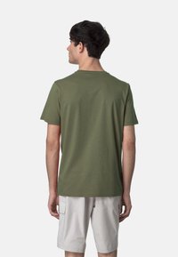 T-shirt verde oliva a maniche corte in cotone testurizzato, con collo a girocollo e vestibilità rilassata. Indossato con pantaloni corti di colore chiaro.