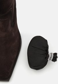 Bruine suede enkellaars met een vierkante neus, textuur oppervlak en gedetailleerde stiksels. Een klein, zwart stoffen zakje ligt ernaast.
