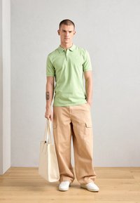 Hellgrünes Poloshirt mit Kragen, kombiniert mit beigen weiten Cargo-Hosen und weißen Sneakers, während er eine cremefarbene Tragetasche hält.