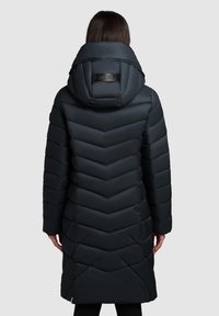 Lange zwarte pufferjas met een chevronpatroon, uitgerust met een grote, gewatteerde capuchon en een merklabel bij de nek.