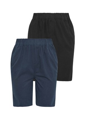 Smilende kvinde med blond hår iført en hvid ærmeløs top, marineblå shorts med elastisk talje og hvide sneakers, der står med den ene hånd i lommen.