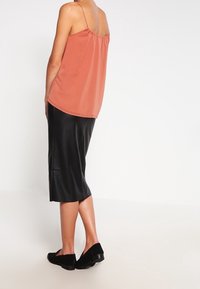 Camisola di raso arancione con spalline sottili, abbinata a una gonna in pelle nera di lunghezza media e scarpe nere senza laccio, vista da dietro.