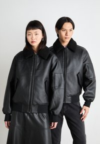 Μαύρες δερμάτινες bomber τζάκετ με γούνινες γιακαδες; διαθέτουν φερμουάρ μπροστά, ριμπ μανσέτες και στρίφωμα, και πλαϊνές τσέπες; δύο μοντέλα στέκοντ αι δίπλα-δίπλα.