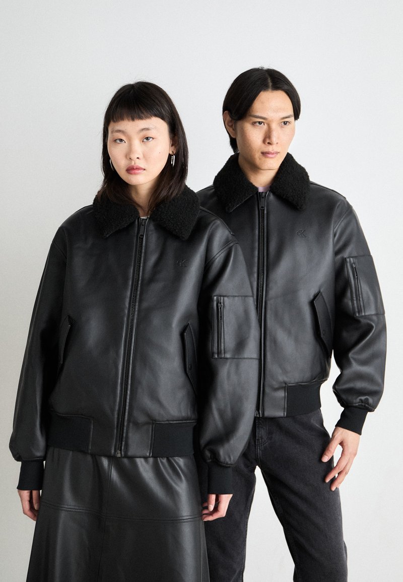 Μαύρες δερμάτινες bomber τζάκετ με γούνινες γιακαδες; διαθέτουν φερμουάρ μπροστά, ριμπ μανσέτες και στρίφωμα, και πλαϊνές τσέπες; δύο μοντέλα στέκονται δίπλα-δίπλα.