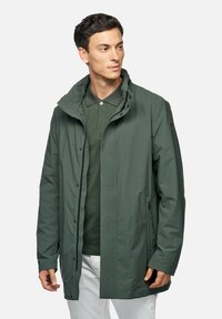 Giubbotto impermeabile verde scuro con cappuccio, chiusure a zip e bottoni a pressione, e tasche laterali inclinate. Tessuto liscio, taglio di lunghezza media.