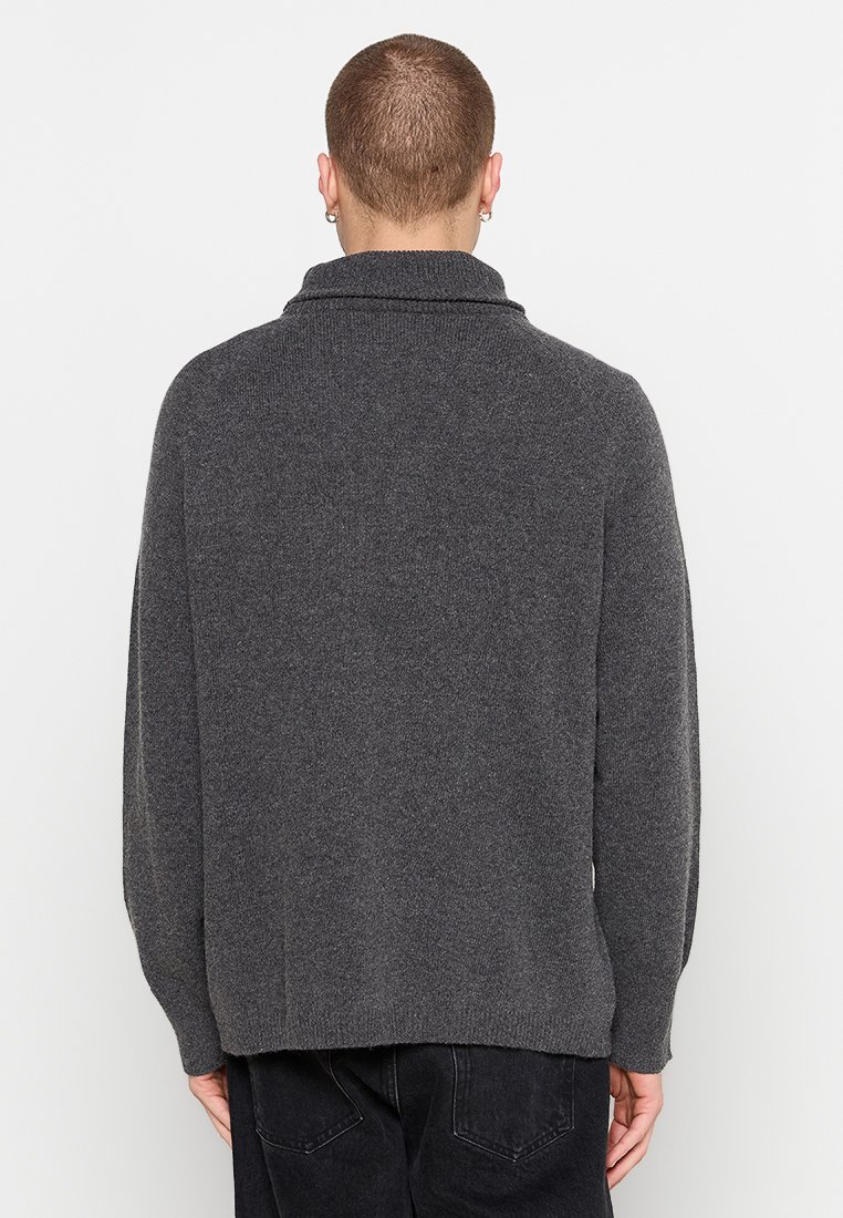 Mann trägt einen dunkelgrauen Langarmpullover und schwarze Jeans, steht mit dem Rücken zu einem schlichten weißen Hintergrund.