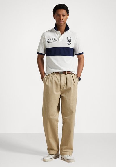 Witte polo met marineblauwe strepen, geborduurd logo en opgestikte kraag; gecombineerd met kaki broek en lichte schoenen, staande tegen een neutrale achtergrond.