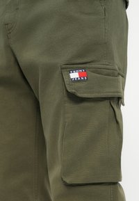 Cargo παντελόνια σε ελαιόλευκο ύφασμα, με πλευρικές τσέπες, ορατή επένδυση λογότυπου με μπλε, κόκκινες και λευκές λεπτομέρειες, και απαλή υφή.