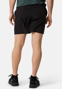 Svarta sportshorts med en slät yta, med resårmidja och avslappnad passform. Bärs tillsammans med svarta skor och strumpor vid vristerna.