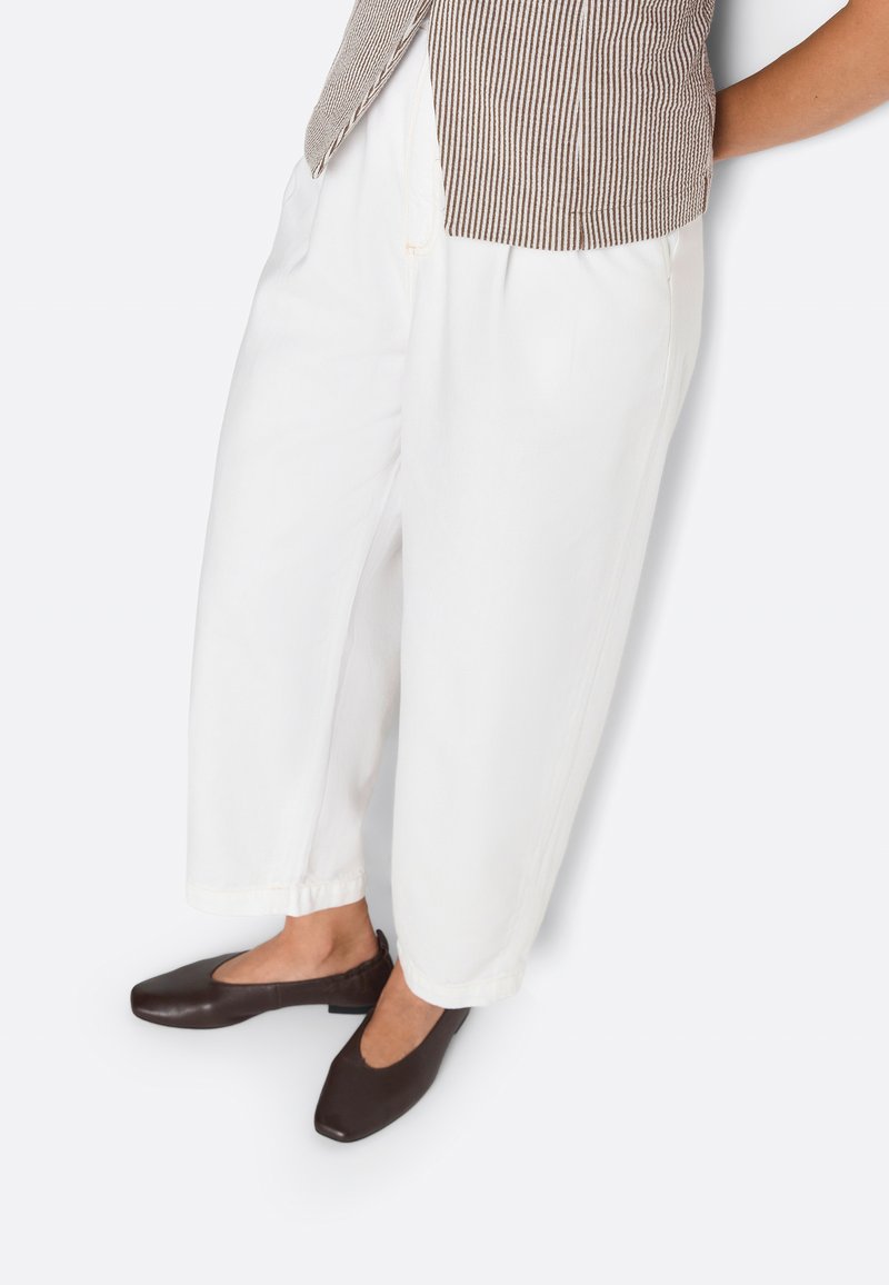 Osoba nosiaca voľné biele skrátené nohavice, hnedý pruhovaný top a tmavohnedé ploché topánky, stojaca pred bielym pozadím.