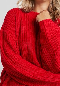 Czerwony sweter z dzianiny o luźnym, teksturowanym wzorze i szerokimi mankietami. Okrągły dekolt i opadające ramiona podkreślają swobodny krój.