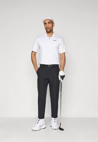 Λευκό πόλο μπλουζάκι με κοντά μανίκια και λογότυπο Nike, μαύρο παντελόνι προσαρμοσμένης γραμμής, λευκά παπούτσια και καπέλο. Κρατάει ένα γκολφ μπαστούνι. Απλός και λειτουργικός σχεδιασμός.