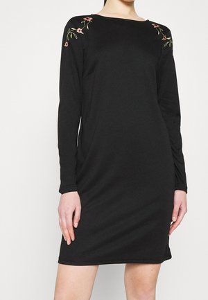 Zwarte jurk met lange mouwen en geborduurde bloemen op de schouders, gedragen door een persoon met ontspannen handen langs de zijden tegen een effen achtergrond.