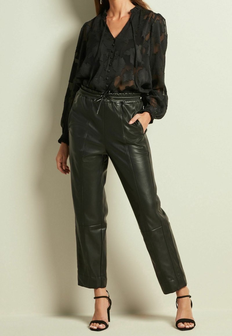 Femme portant un chemisier noir transparent à fleurs avec de longues manches bouffantes, associé à un pantalon en cuir noir taille haute et des talons noirs à brides.