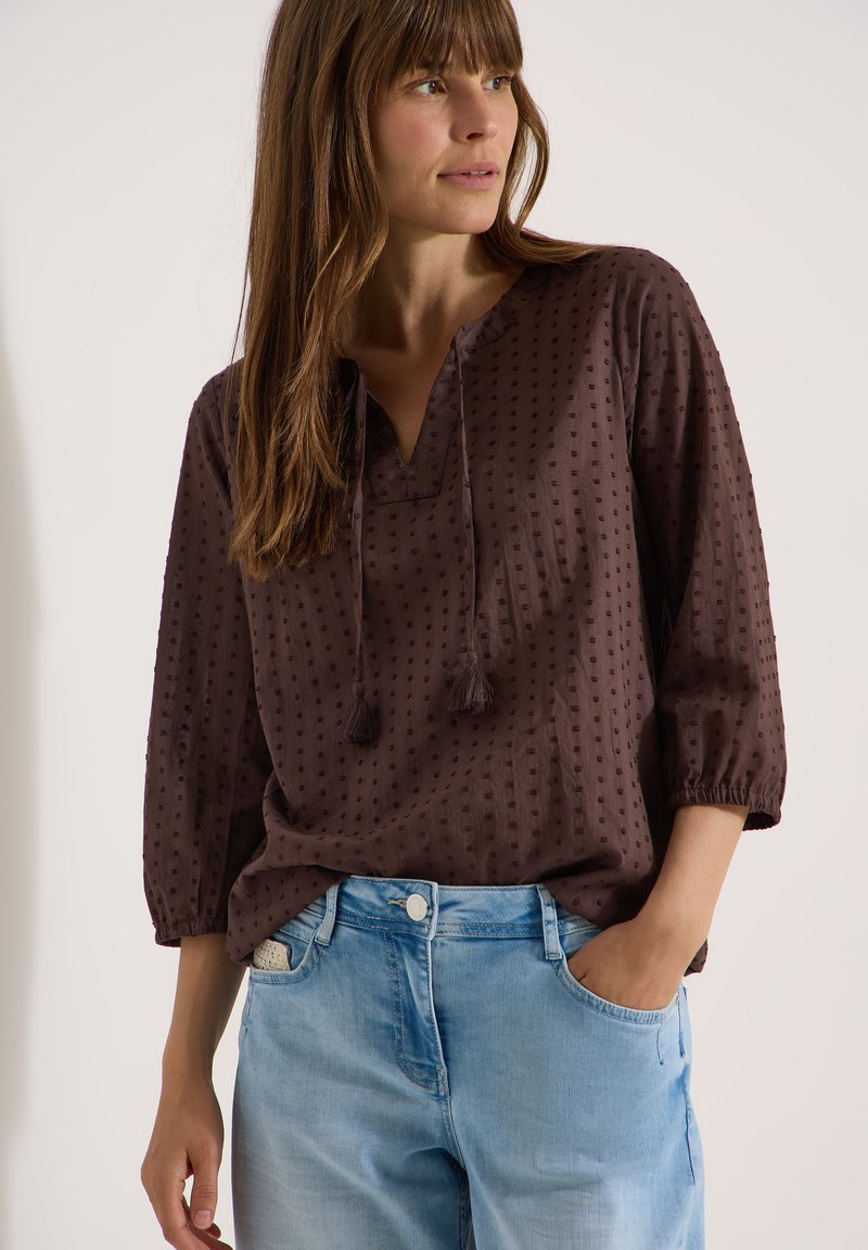 Femme portant un chemisier texturé marron foncé avec des liens à pompons et un jean en denim bleu clair, regardant sur le côté avec une main dans la poche.
