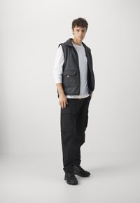Grå quilted vest med lynlås og to frontlommer, lagd over en hvid langærmet skjorte, parret med sorte cargo-bukser og sneakers.