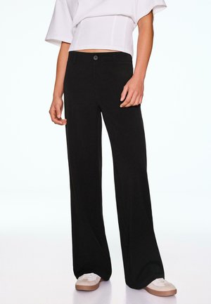 Personne portant un pantalon noir à jambes larges, un top blanc court et des baskets blanches, debout devant un fond clair uni.